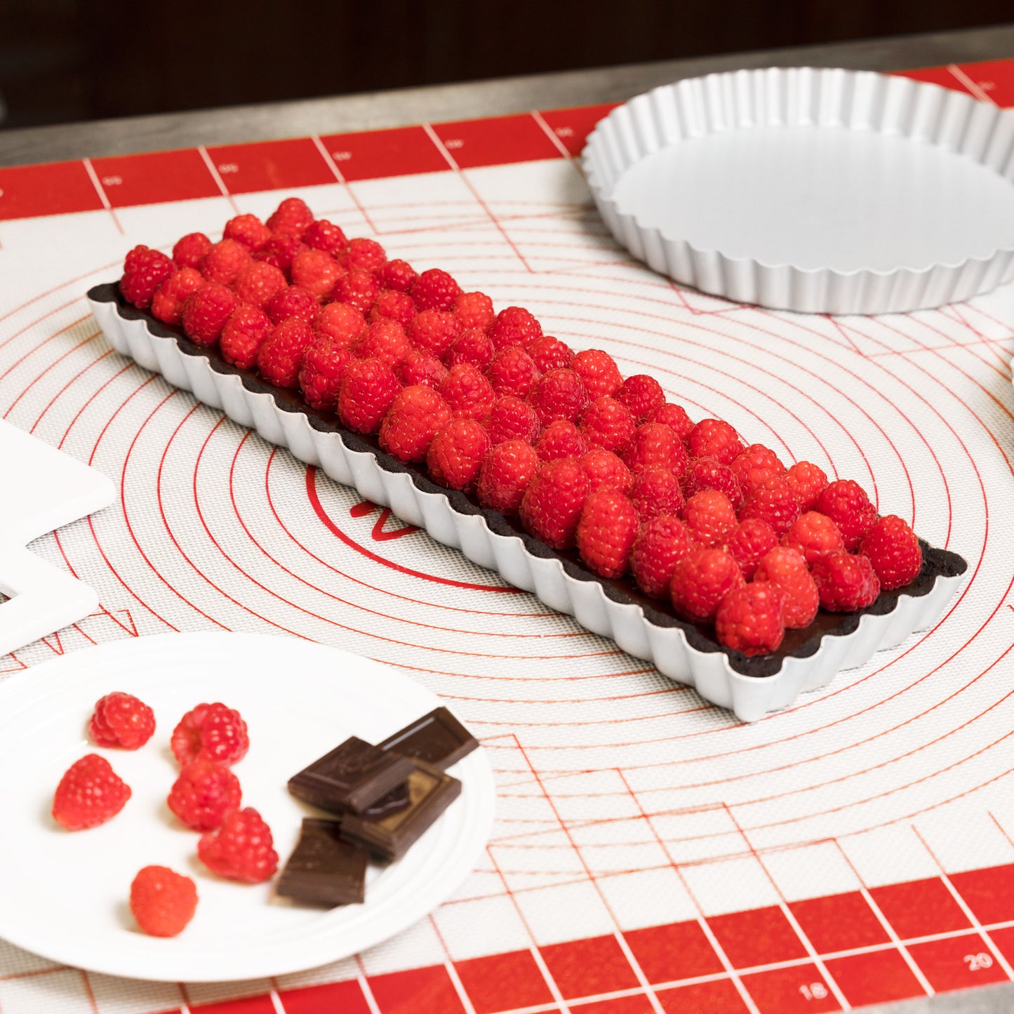 Rectangle chocolate raspberry tart in a tart and quiche pan with raspberries and chocolate on a silicone baking mat in a kitchen