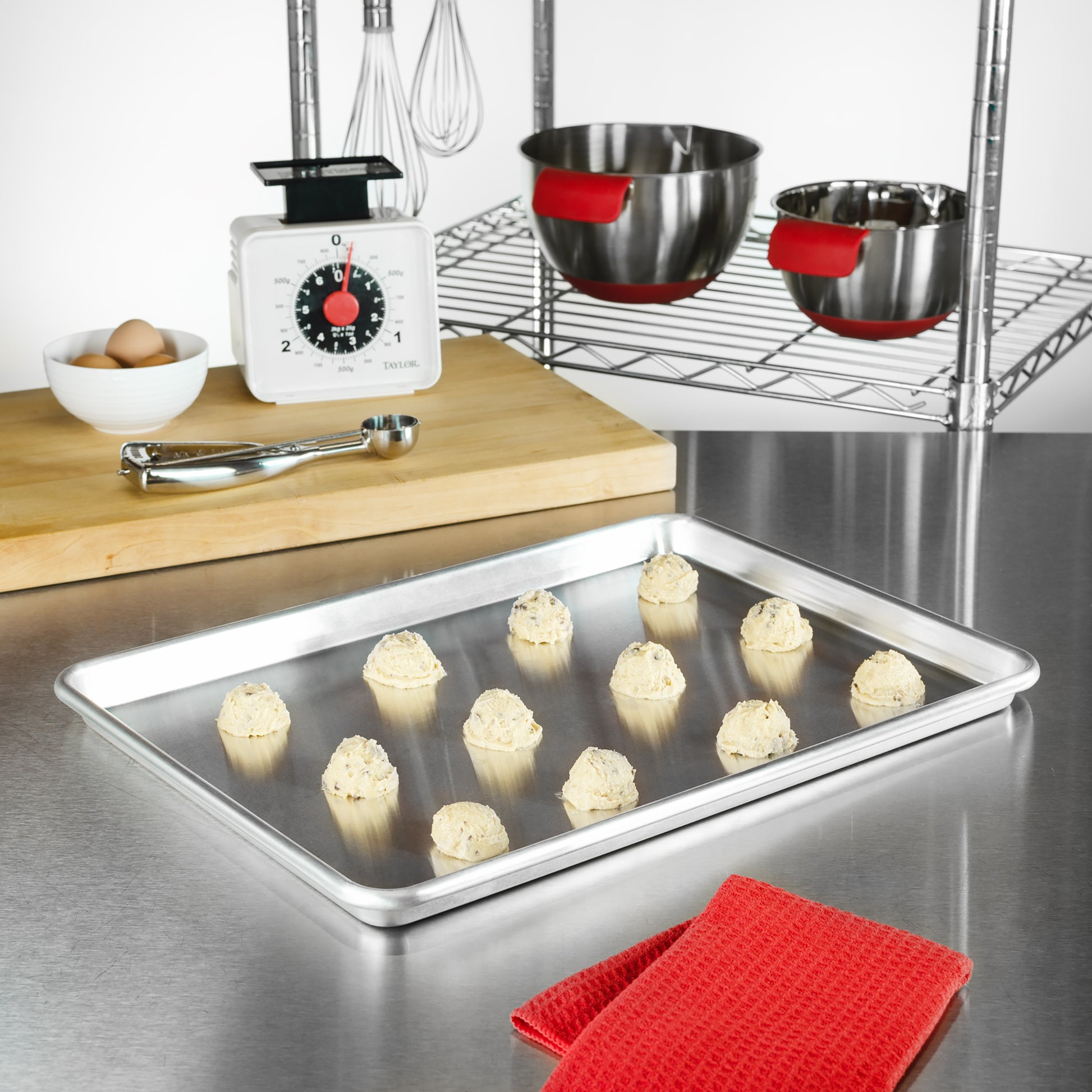 Half baking sheet pan with cookie dough, eggs, cookie scoops, kitchen scale, tools and bowls in a bakery